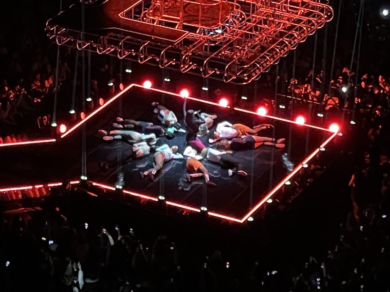 Dua Lipa on a diamond-shaped stage, kneeling, surrounded by dancers